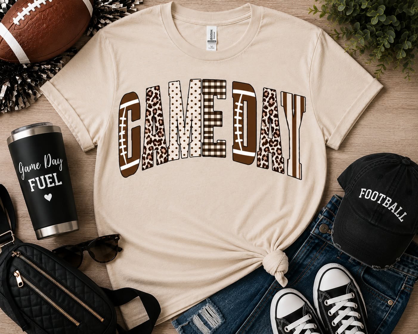 A beige graphic T-shirt laid flat on a wood surface with football-themed props including a football, cap, sunglasses, tumbler, jeans, and shoes. The shirt has large arched text reading “GAME DAY” in mixed brown patterned blocks.