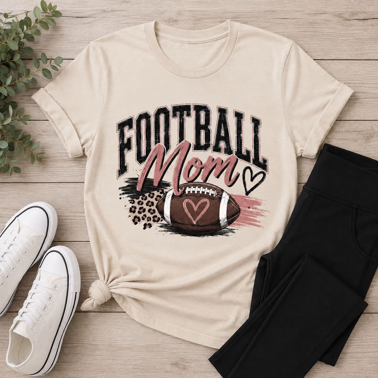 Cream Football Mom graphic T-shirt with distressed black lettering, blush pink accents, leopard print details, and a football with hearts.